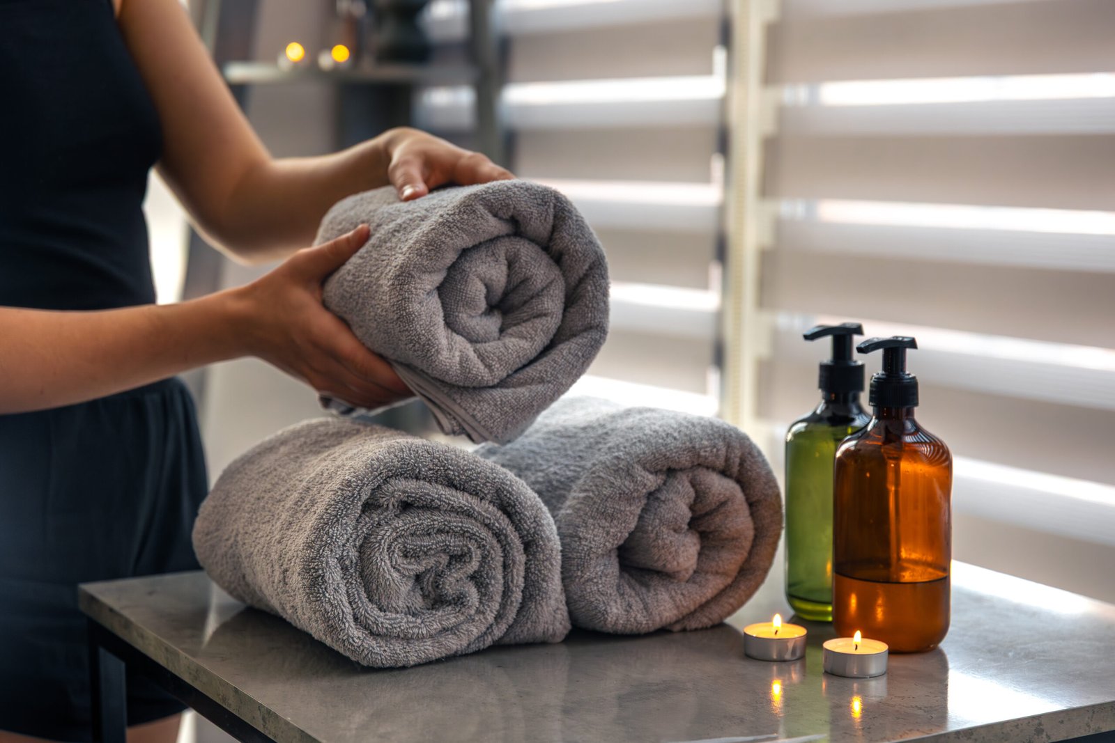 Hands hold a fresh roll above two towels with candles and pump bottles. Quiet minimalist setting for everyday body care.